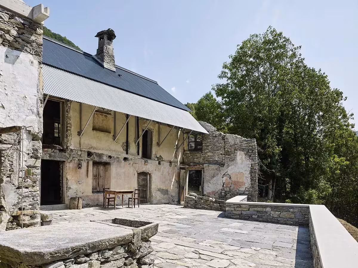 Fachada de Casa Mosogno con cubierta de chapa nueva sobre muros de piedra y la terraza de losas, en el valle de Onsernone