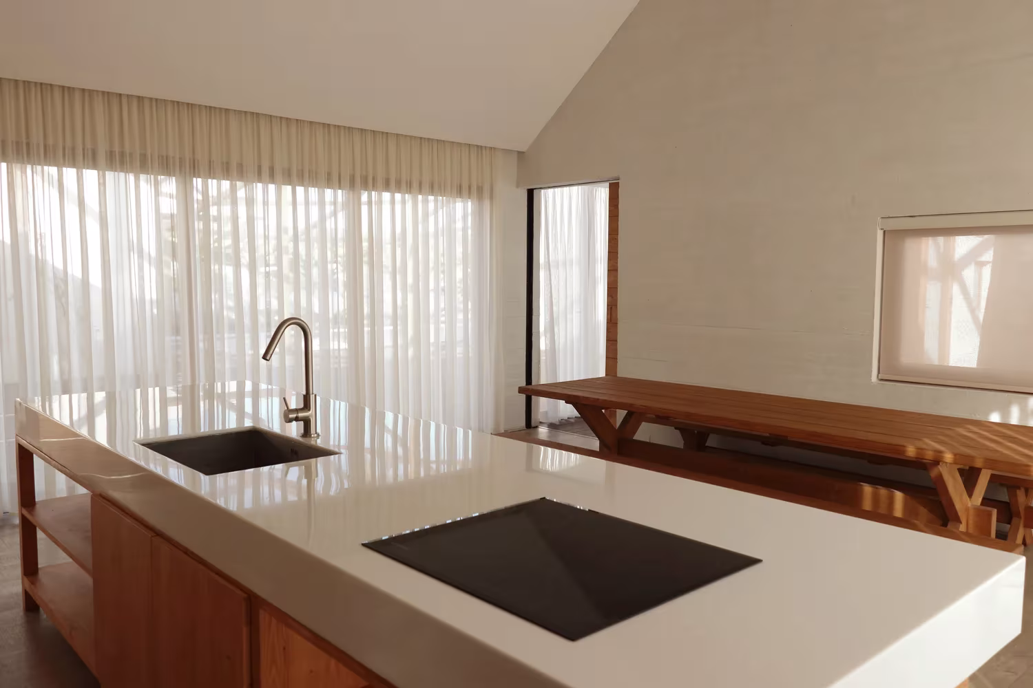 Cocina de Villa Vanoosh con isla blanca y cortinas de visillo ante un gran ventanal, junto a una mesa de madera