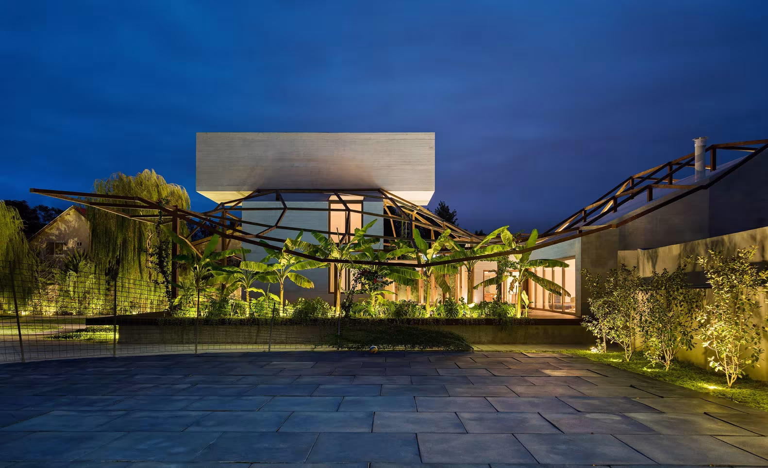 Villa Vanoosh iluminada al anochecer, con la plaza de piedra, la vegetación y el volumen elevado