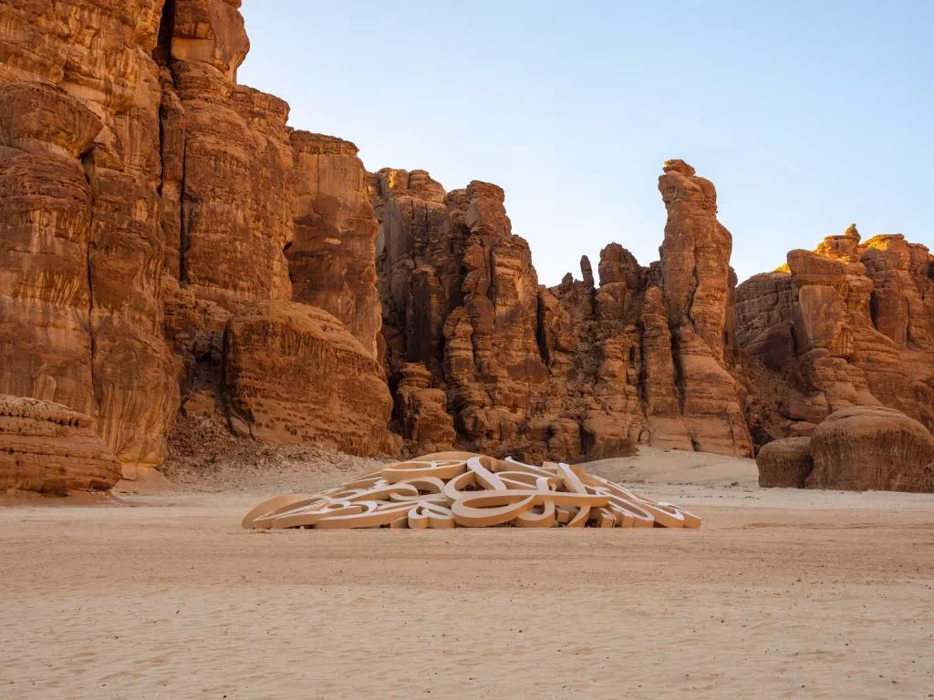 Escultura de tubos entrelazados color arena sobre el suelo del desierto de AlUla, entre acantilados de arenisca