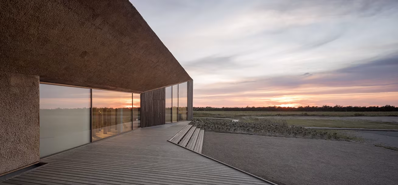Voladizo de la cubierta de paja y fachada acristalada sobre la tarima, con la puesta de sol sobre el Mar de Wadden