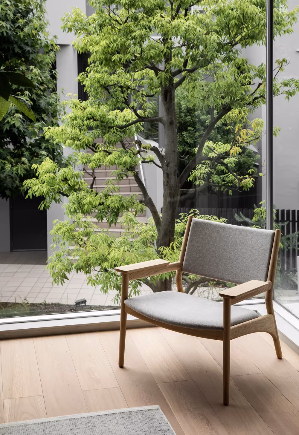 Butaca de madera y tapizado gris junto a un ventanal con un arce en el patio de Kinuta Terrace, Tokio