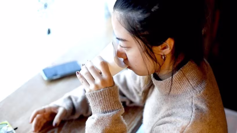 Mujer bebiendo de un vaso de papel Unocup con tapa plegable, sentada a una mesa junto a un móvil