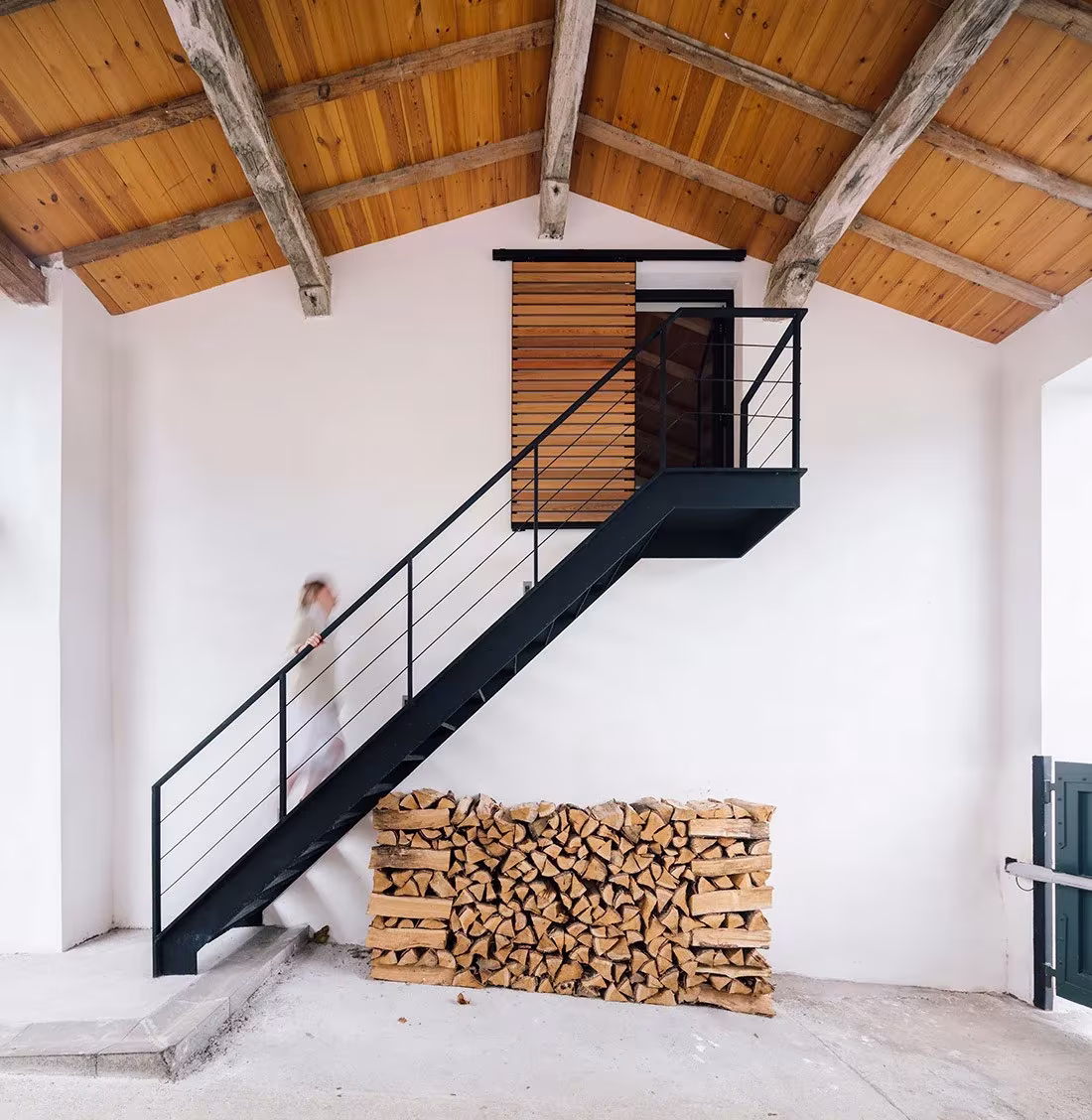 Escalera metálica negra y leñera apilada bajo el faldón de madera en la casa de Zooco en Güemes