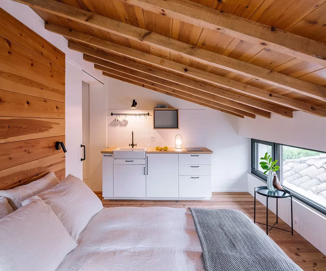 Dormitorio abuhardillado con techo de madera, cocina pequeña y ventana corrida en la casa de Zooco, Güemes