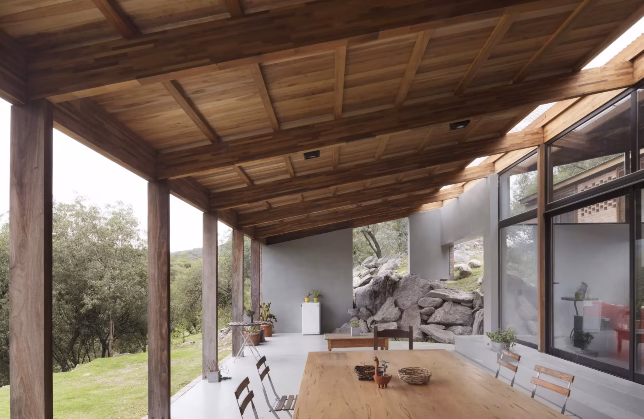 Galería de la Casa JB con pilares de madera, mesa larga y una roca natural dentro de la vivienda