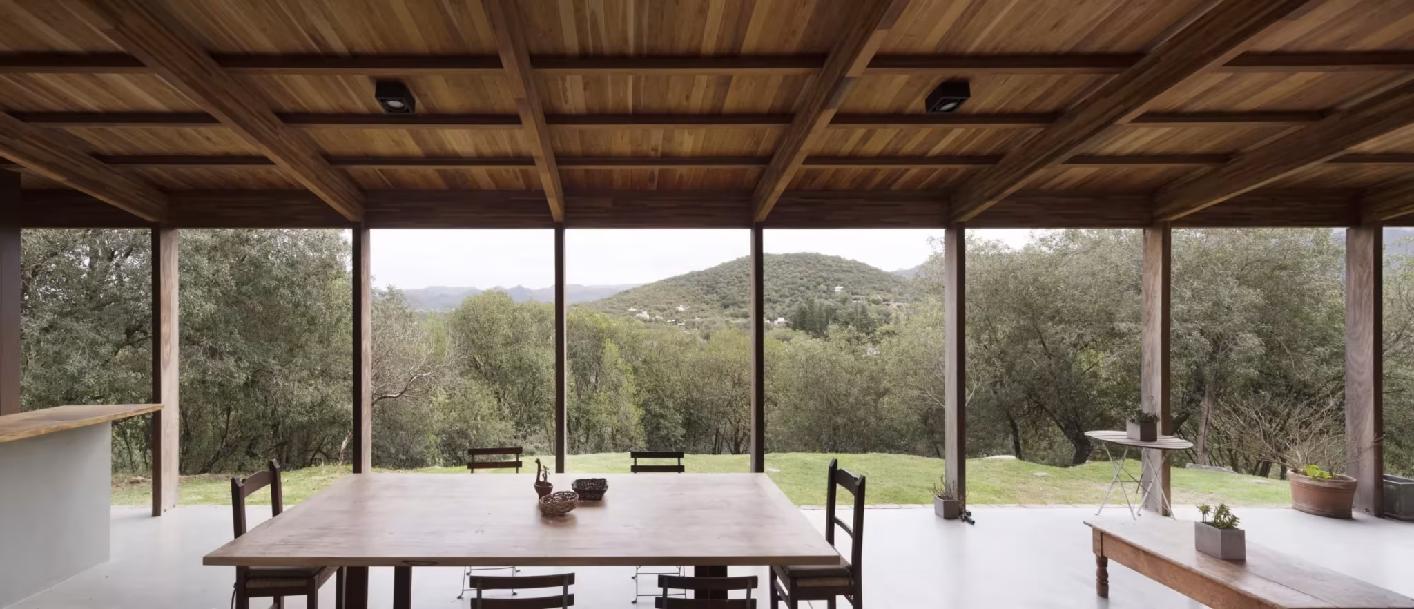 Comedor con mesa de madera bajo techo de vigas, abierto al paisaje serrano de la Casa JB en Unquillo