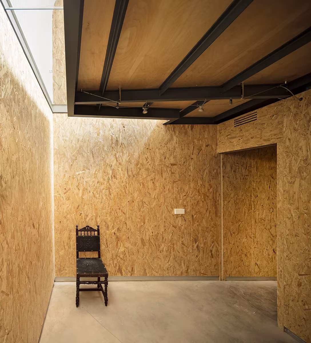 Sala de paredes de OSB con vigas metálicas y luz cenital sobre una silla de madera en Casa Piedrabuena