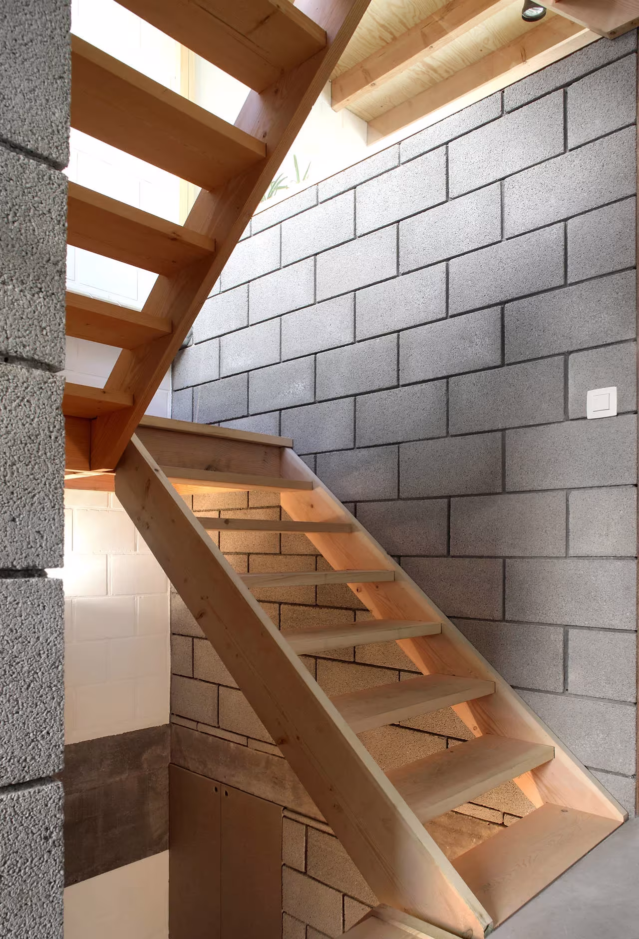 Tramo de escalera de madera junto a muros de bloque de hormigón con luz cenital en la Casa 12k