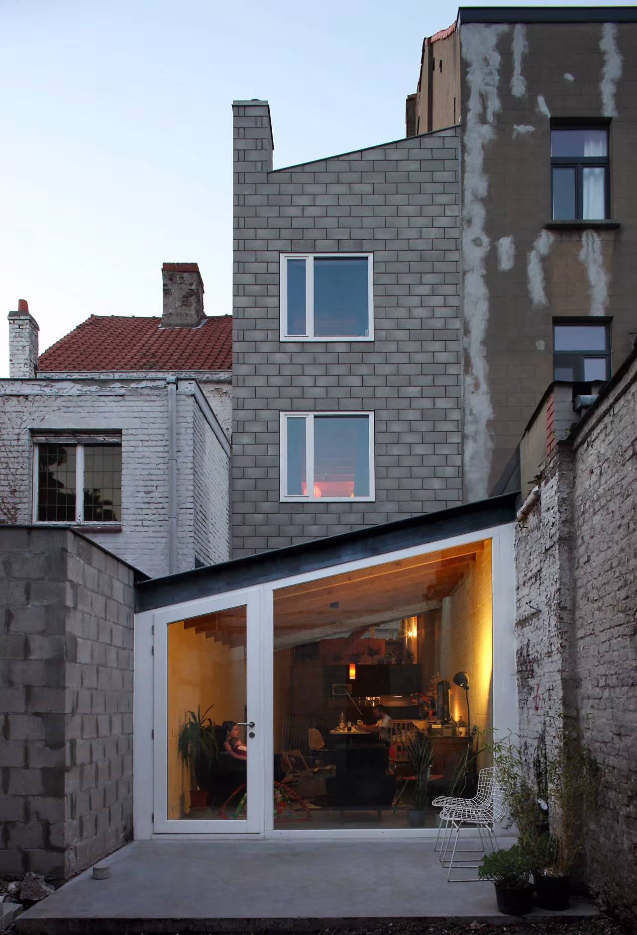 Fachada trasera de bloque gris con volumen acristalado iluminado sobre el patio de la Casa 12k