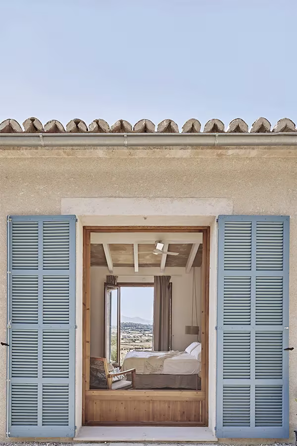Casa Palerm de OHLAB: puerta abierta con contraventanas azules que deja ver un dormitorio y el campo mallorquín