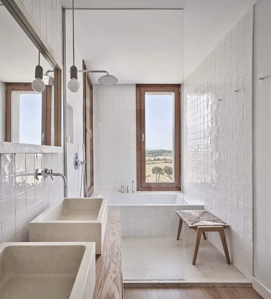 Baño de la Casa Palerm con azulejo blanco, lavabos de piedra, bañera, banco de madera y ventana al campo
