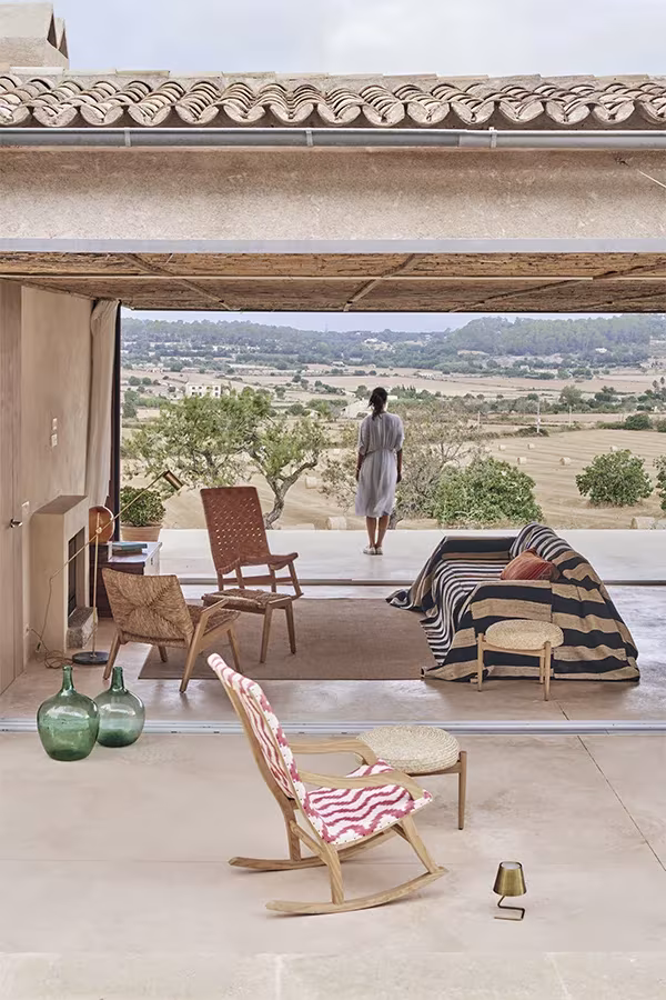 Porche de la Casa Palerm abierto al paisaje rural, con mecedora de rayas, butacas y una persona al fondo