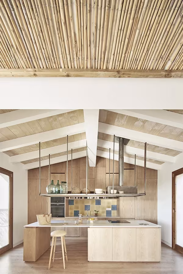Cocina de la Casa Palerm con isla de madera clara, techo de vigas y cañizo, y campana colgante de acero