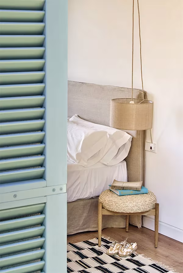 Dormitorio de la Casa Palerm visto tras una contraventana verde, con cama blanca, lámpara colgante y taburete