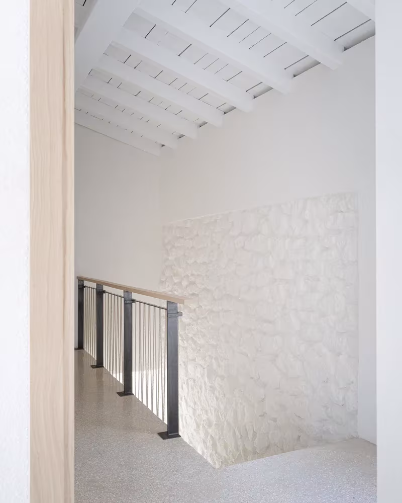 Hueco de escalera con barandilla metálica y muro de piedra encalada bajo techo de vigas blancas en la Casa MA