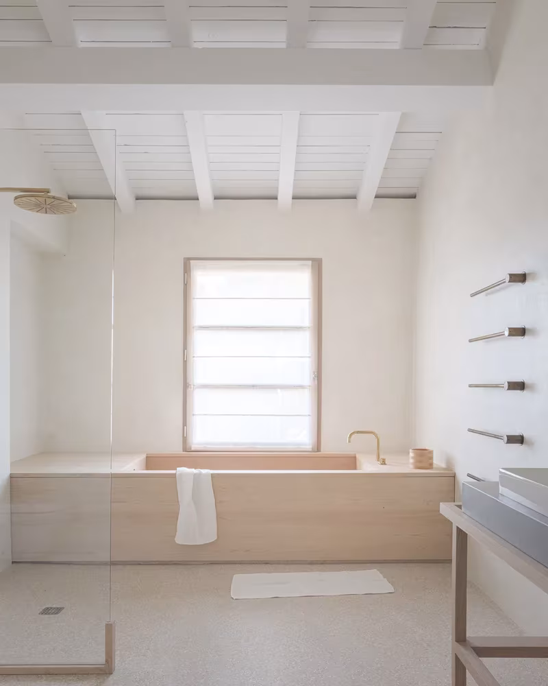 Baño de la Casa MA con bañera de madera, ventana pequeña y mampara de vidrio bajo techo de vigas blancas