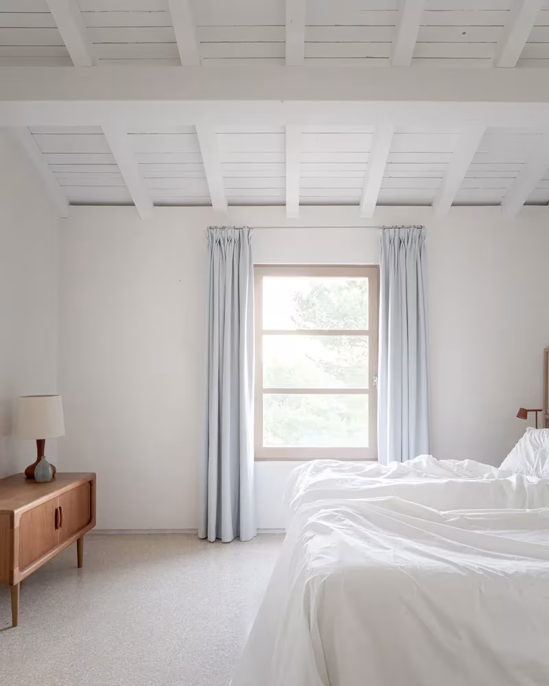 Dormitorio blanco de la Casa MA con ventana de marco de madera, cortinas azul pálido y cómoda de madera