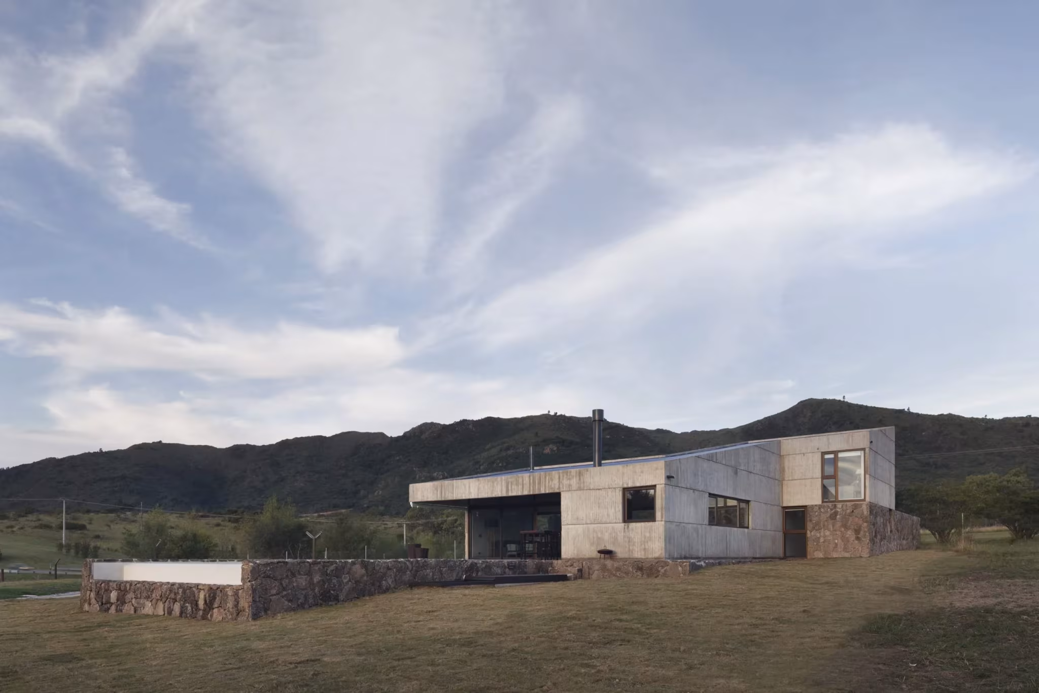 Casa MM con zócalo de piedra y volúmenes de hormigón frente a las montañas de Calamuchita