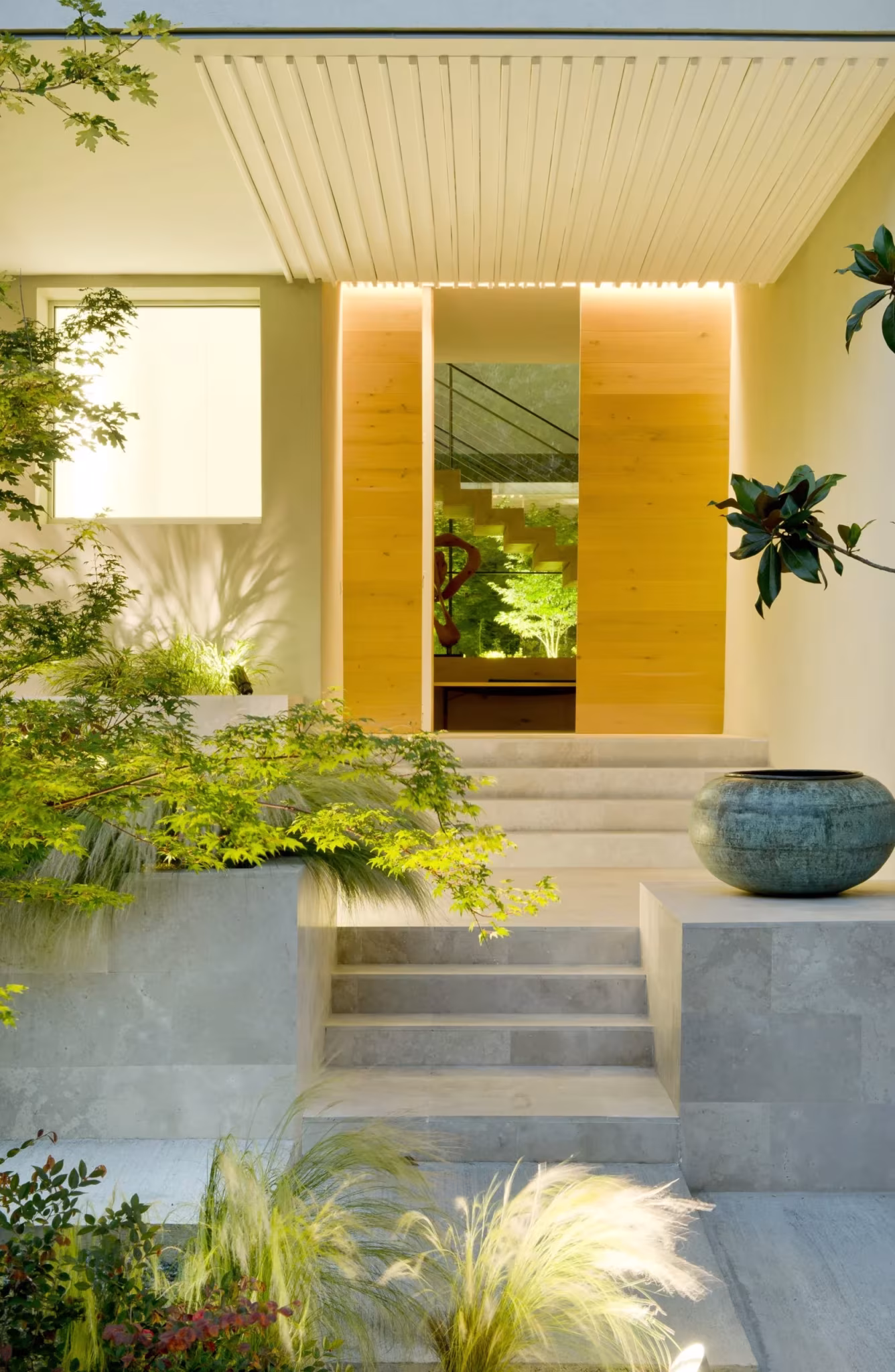 Entrada con pérgola de lamas blancas, panel de madera iluminado y escalones entre plantas de jardín