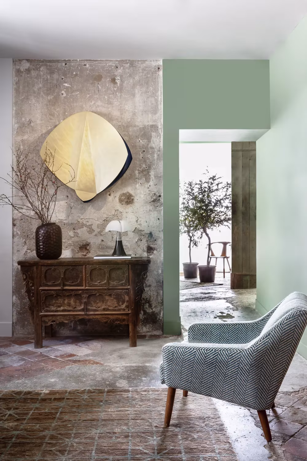 Recibidor con aparador antiguo de madera, espejo dorado, pared verde y butaca estampada sobre solería de barro