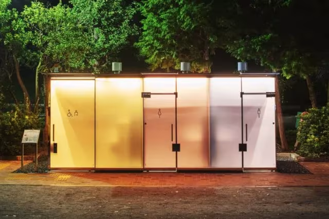 Baños públicos de Shigeru Ban iluminados de noche, con el vidrio opaco brillando como un farol entre árboles