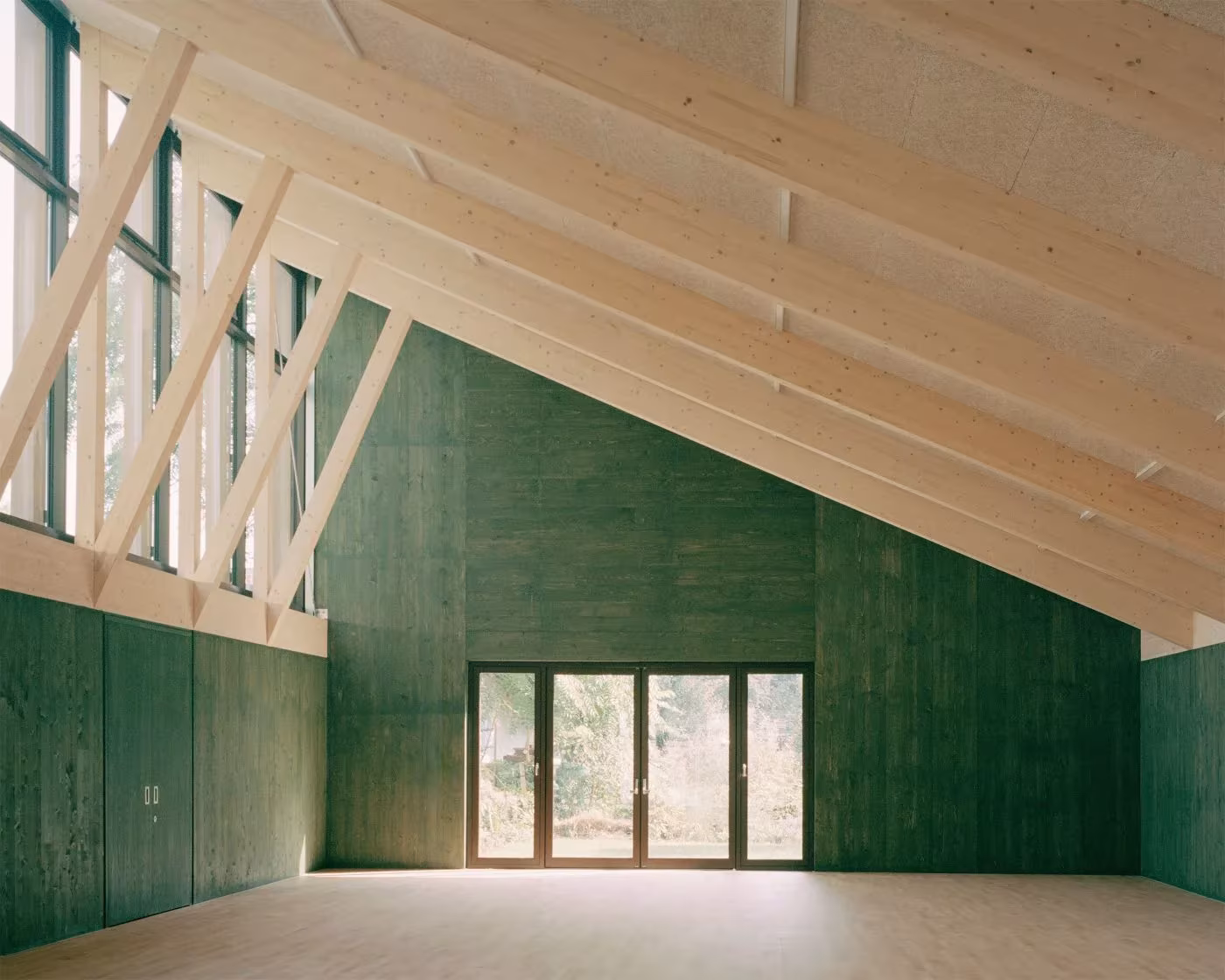Sala del Sands End Centre con cerchas de madera clara, paredes verdes y puertas acristaladas al fondo