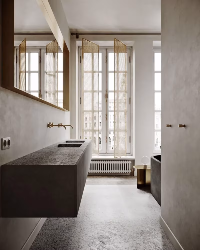 Baño con lavabo de piedra oscura bajo espejo de latón y ventanales con vistas a la calle
