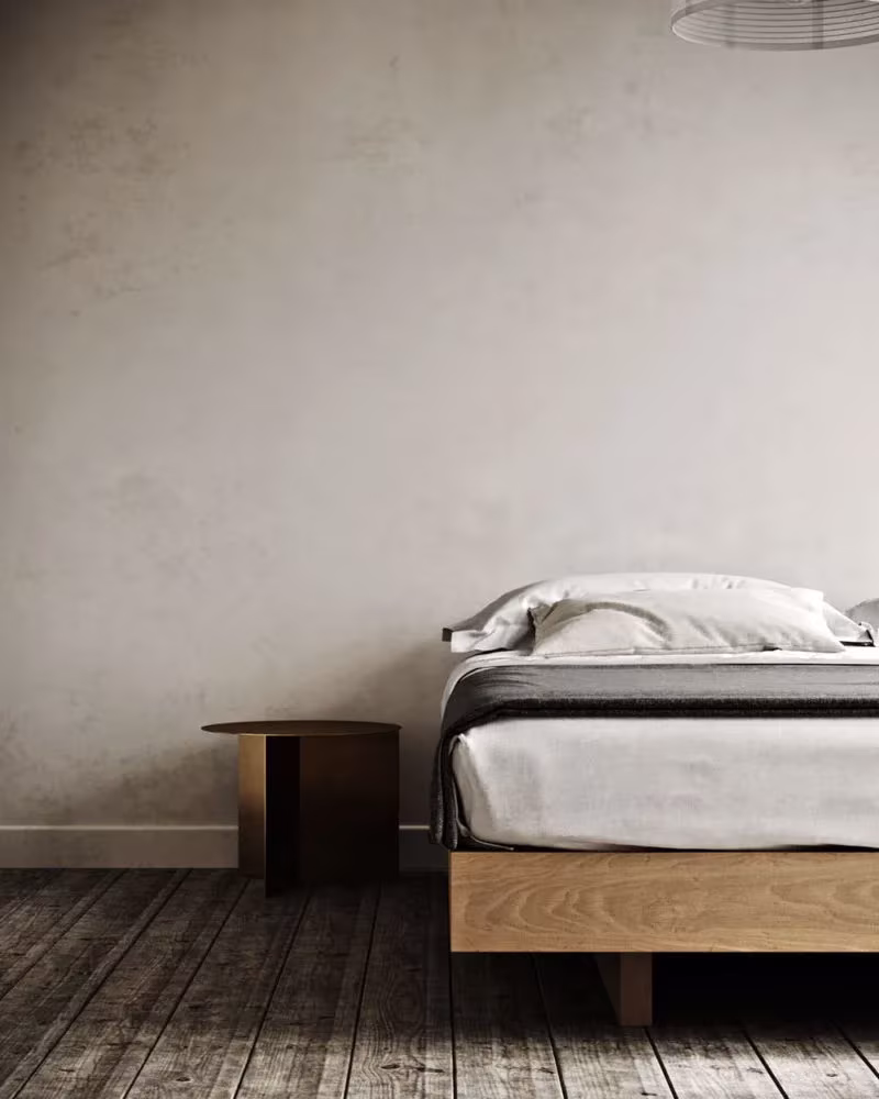 Cama baja con colcha gris sobre base de madera y mesilla redonda en un dormitorio de paredes de estuco