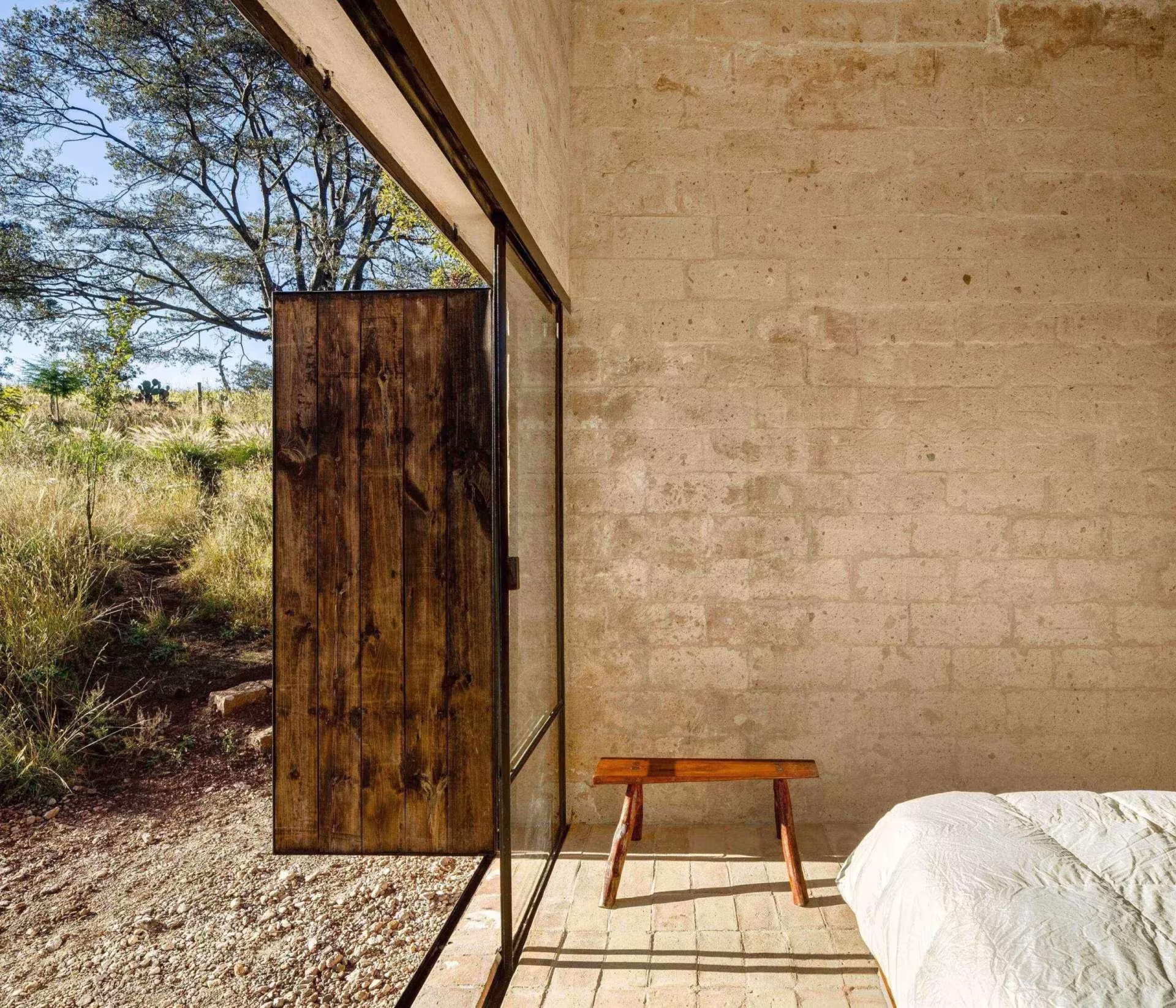 Dormitorio del refugio de Aculco con muro de piedra, banco de madera, cama blanca y puerta corredera al campo