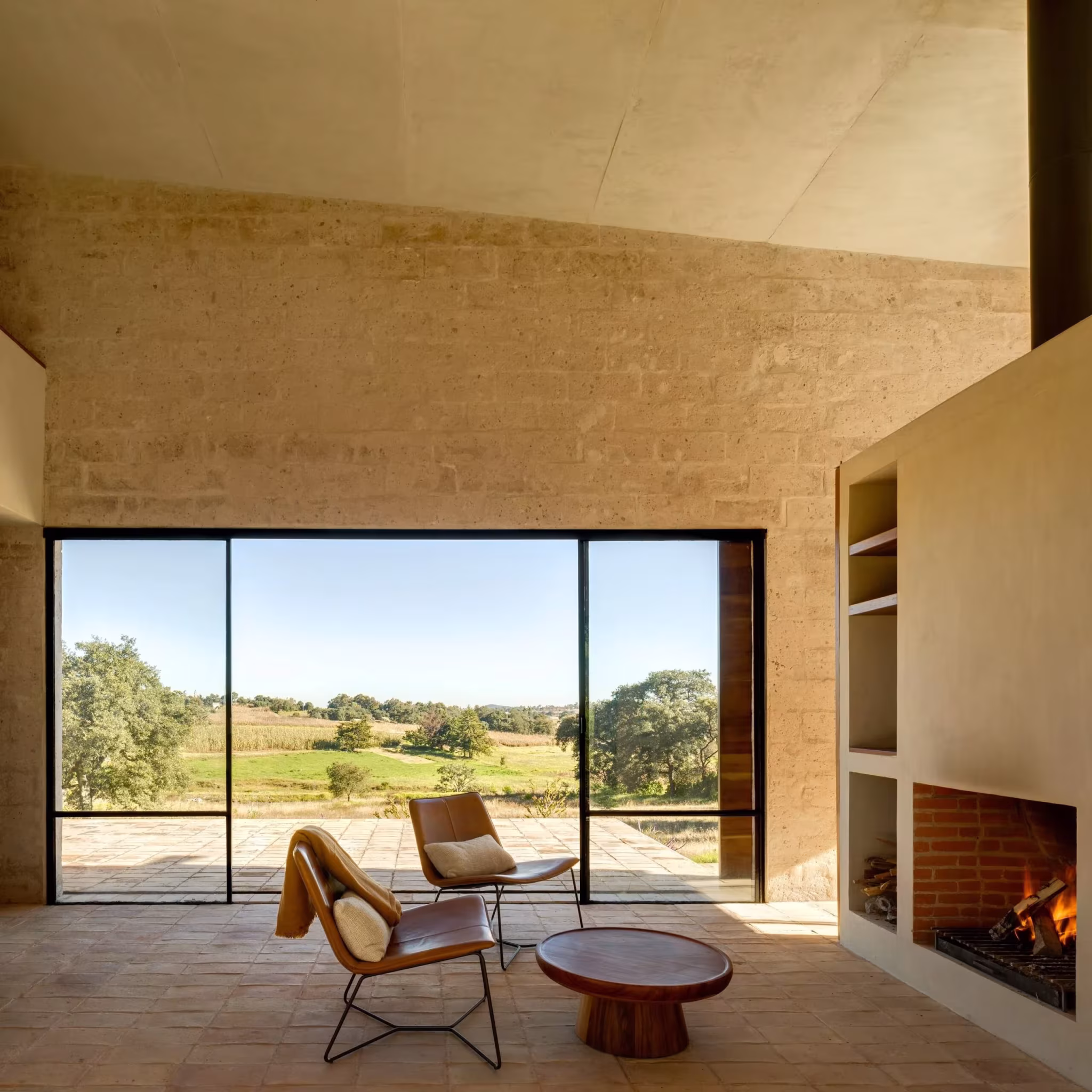 Estancia del refugio de Aculco con chimenea encendida, butacas y ventanal hacia un paisaje de encinas