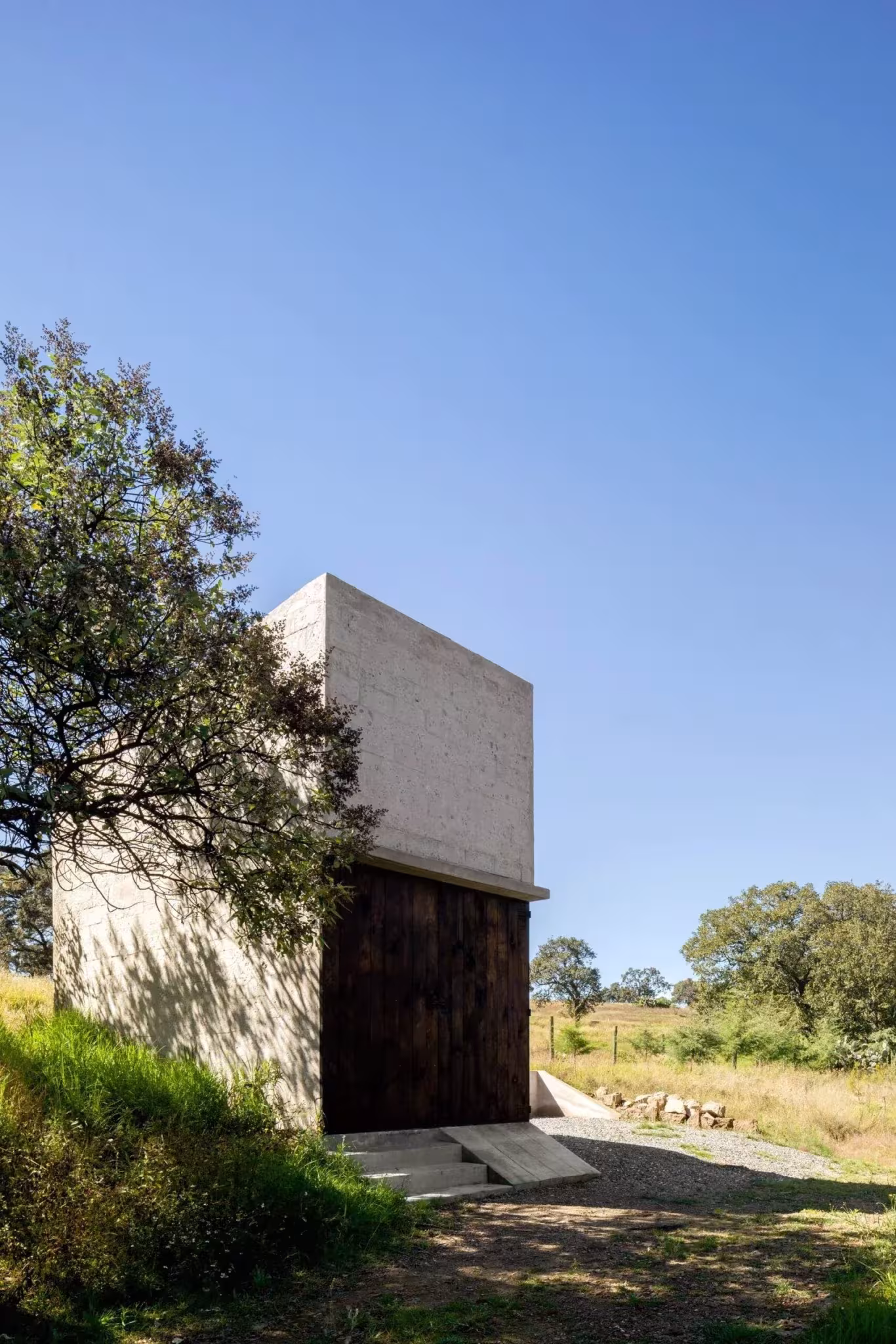 Volumen vertical de piedra del refugio de Aculco con puerta de madera, junto a un gran árbol