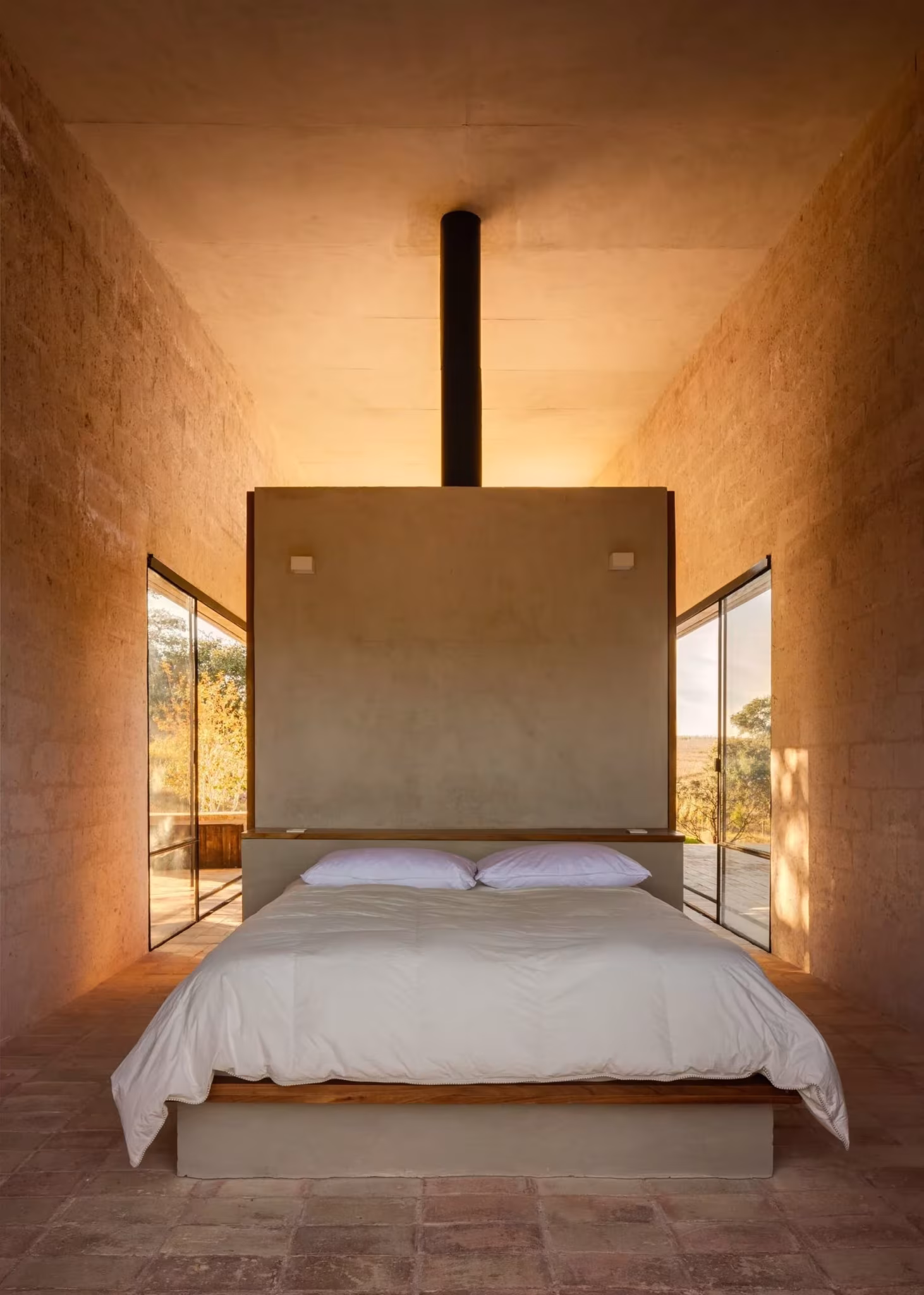 Dormitorio del refugio de Aculco con cama blanca ante un cabecero de piedra y luz cálida del atardecer