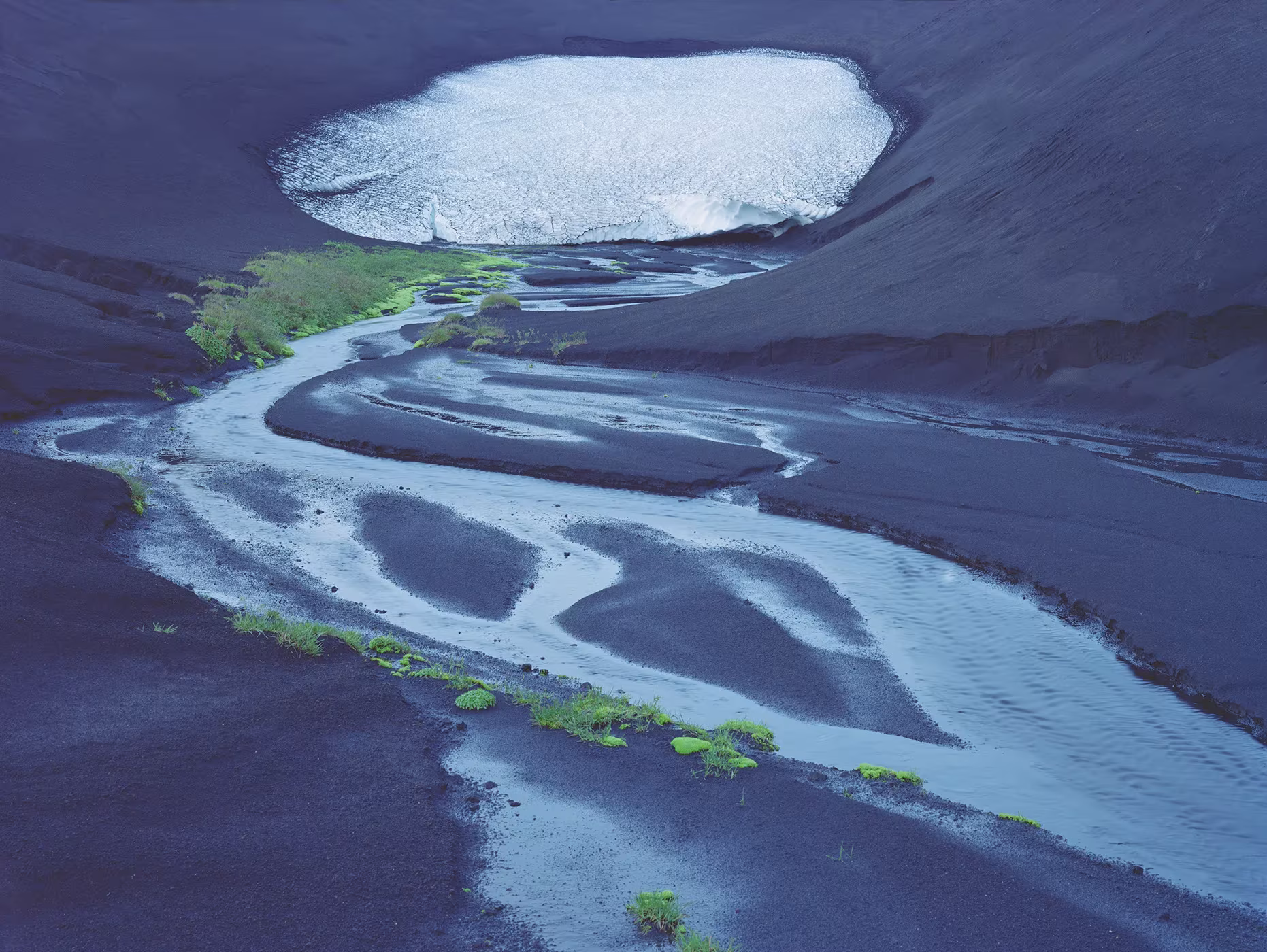 Vista aérea de un río trenzado entre arenas negras junto a una laguna helada, de Luca Tombolini