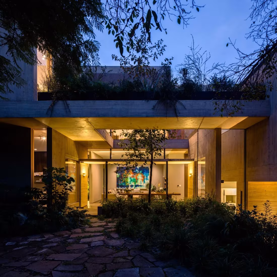Salón de la Casa CBC iluminado al anochecer bajo el voladizo, visto entre vegetación y un camino de piedra
