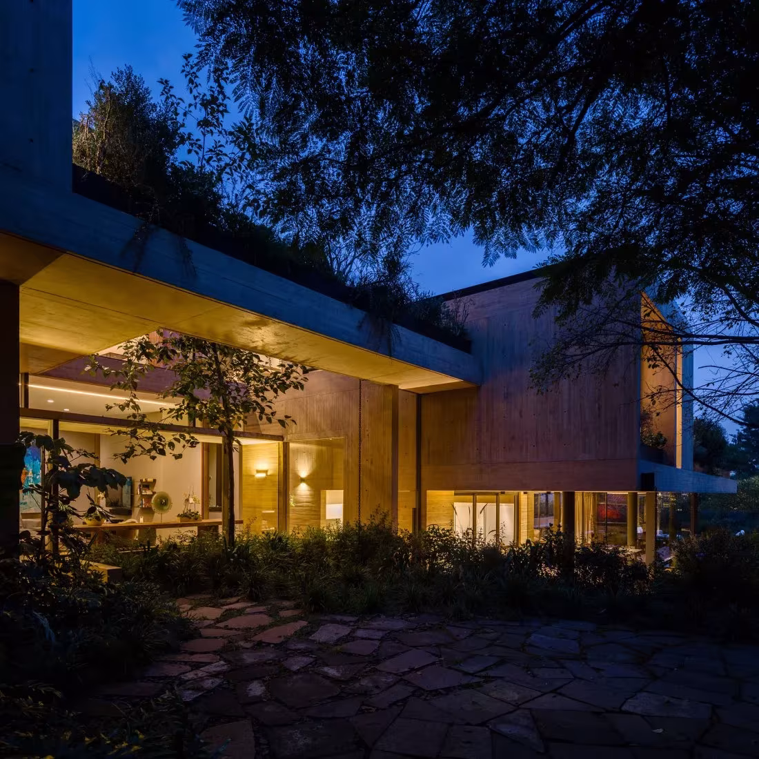 Fachada de la Casa CBC al anochecer, con el salón iluminado tras los árboles y un suelo de losas de piedra