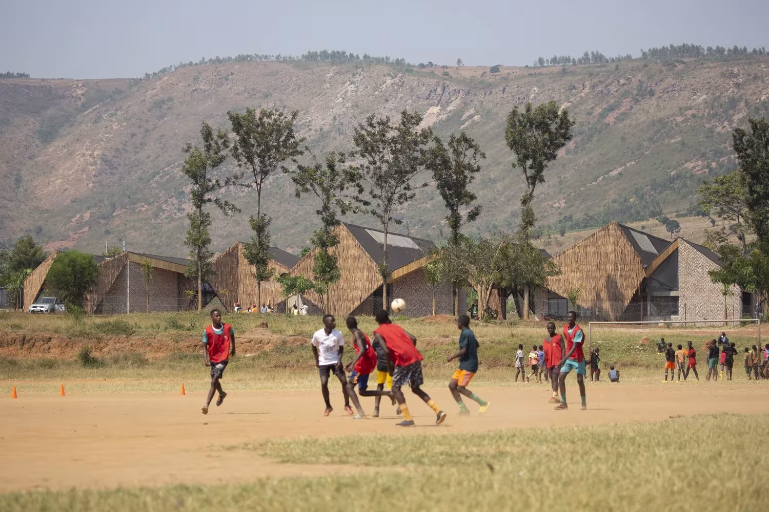 Jóvenes jugando al fútbol en un campo de tierra ante el Centro de Liderazgo Komera y sus cubiertas inclinadas