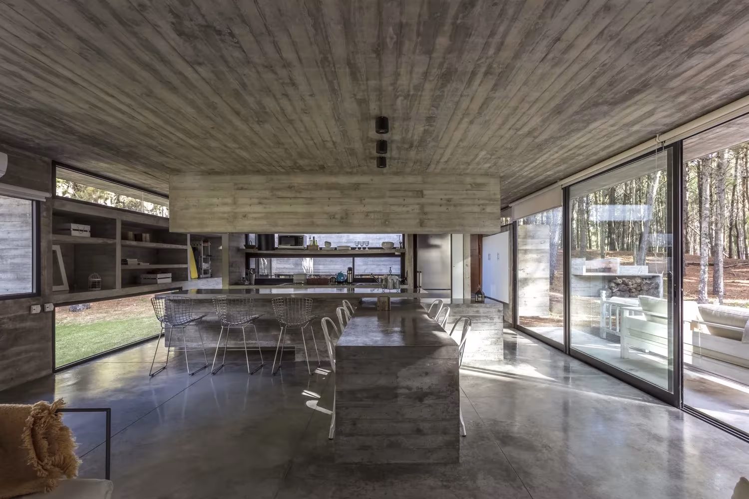 Cocina y comedor de la Casa Bosque con isla de hormigón, sillas blancas y grandes correderas de vidrio