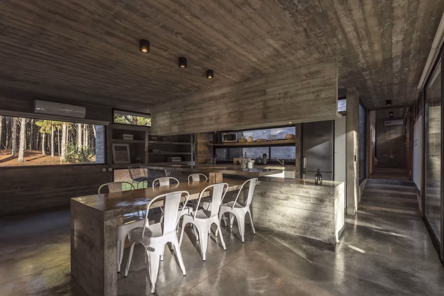 Comedor de la Casa Bosque con mesa de hormigón, sillas metálicas blancas y ventanas horizontales al bosque