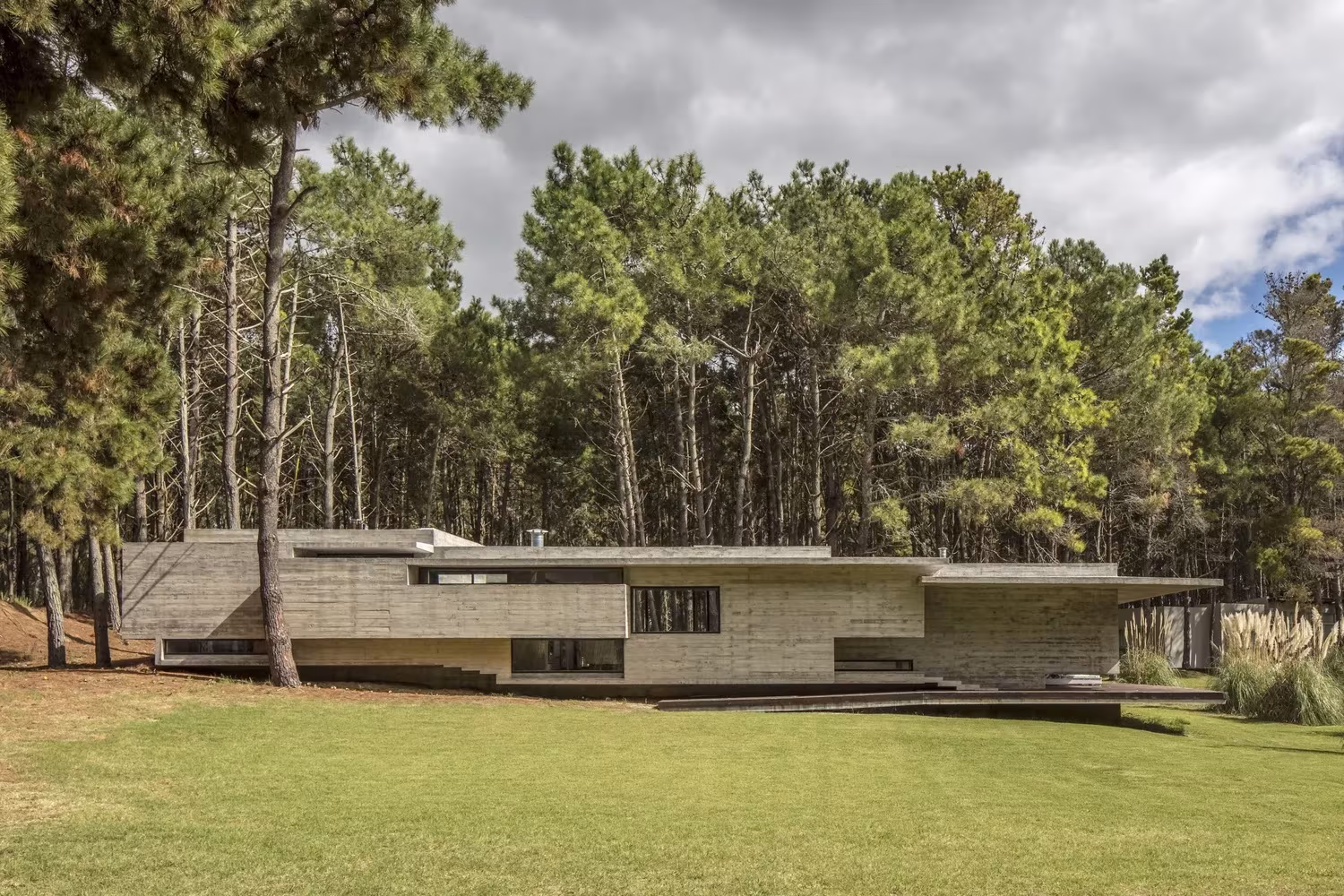 Vista lejana de la Casa Bosque, volumen bajo de hormigón en un claro de césped rodeado de pinos