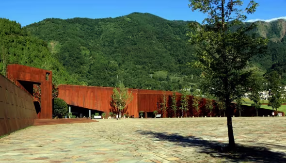 Muro de acero corten junto a una plaza empedrada y montañas verdes en el Museo del Terremoto de Wenchuan