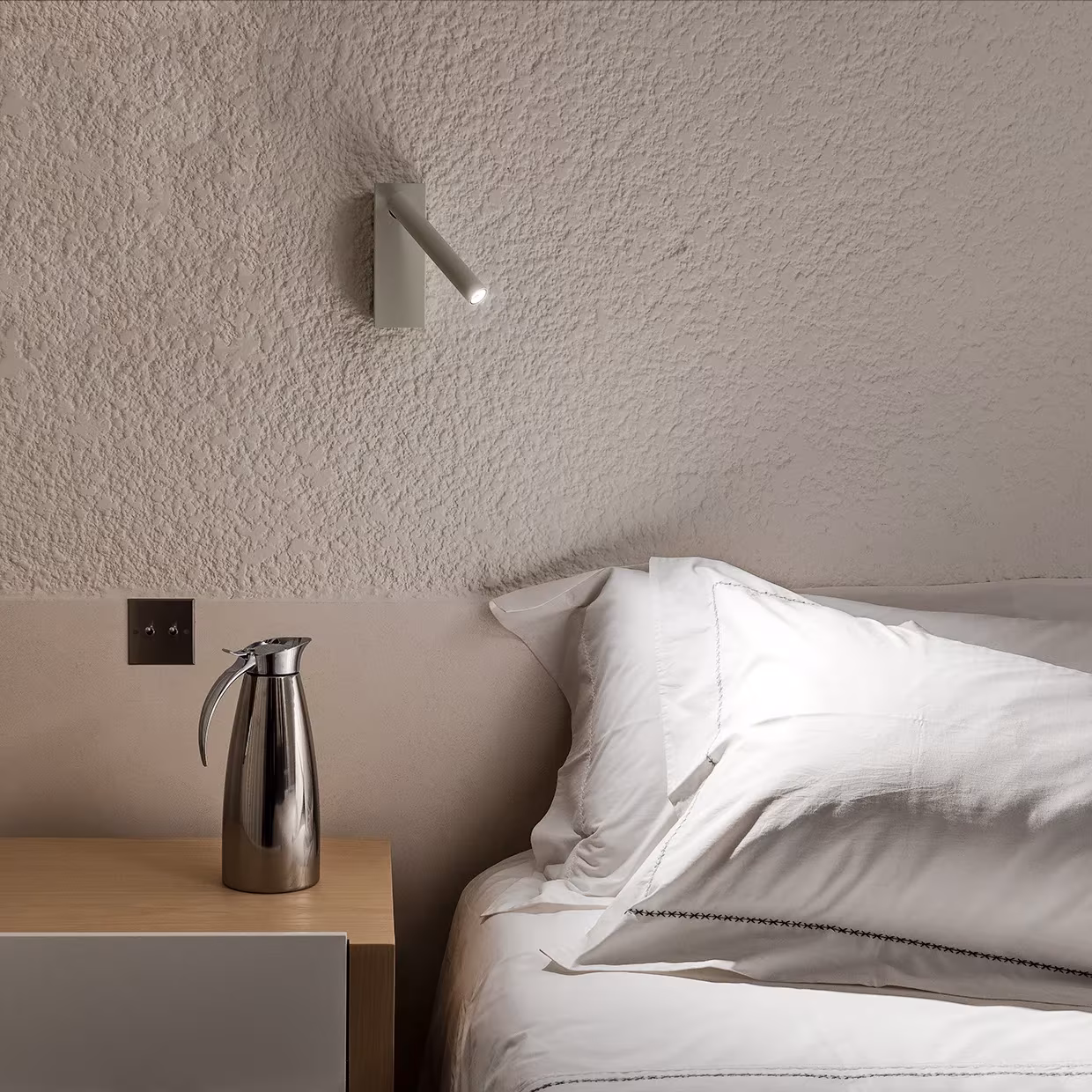Detalle de dormitorio en La Grintosa con cama de sábanas blancas, termo metálico y aplique de lectura en pared rugosa