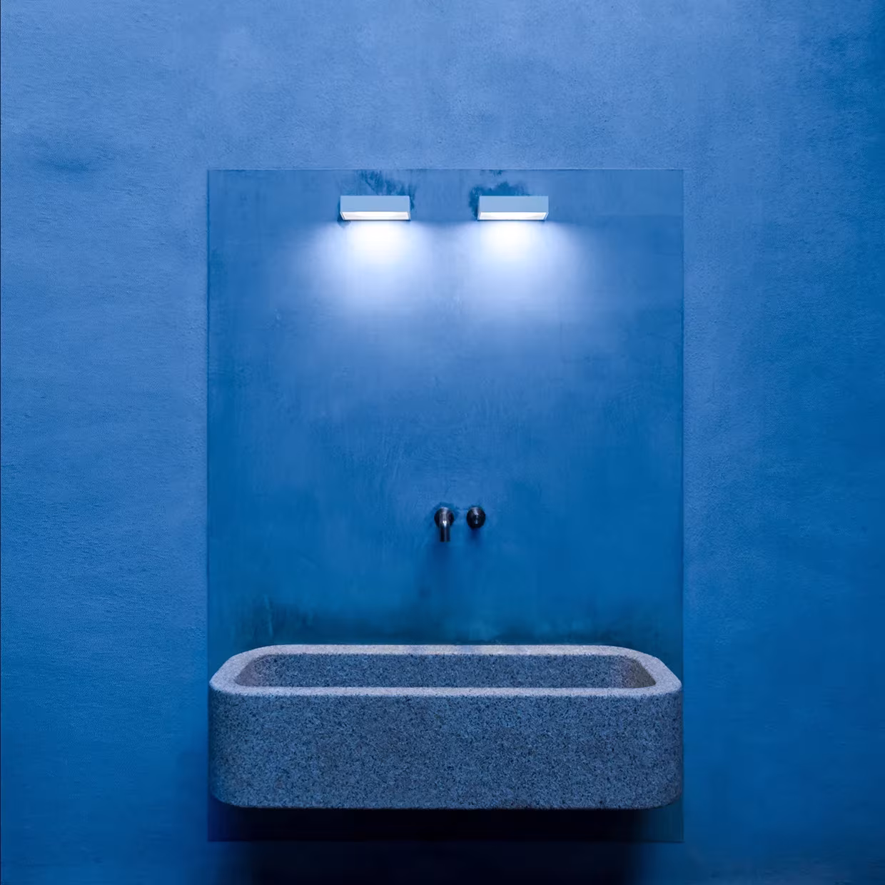 Lavabo de piedra maciza con dos apliques sobre una pared azul en La Grintosa de Stefania Stera