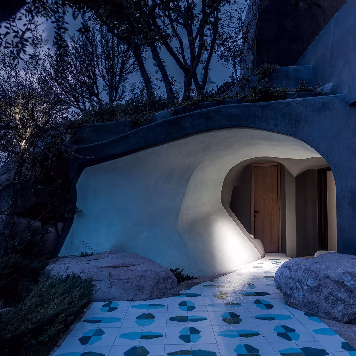 Entrada cueva de formas curvas blancas con puerta de madera y suelo de baldosas azules en La Grintosa