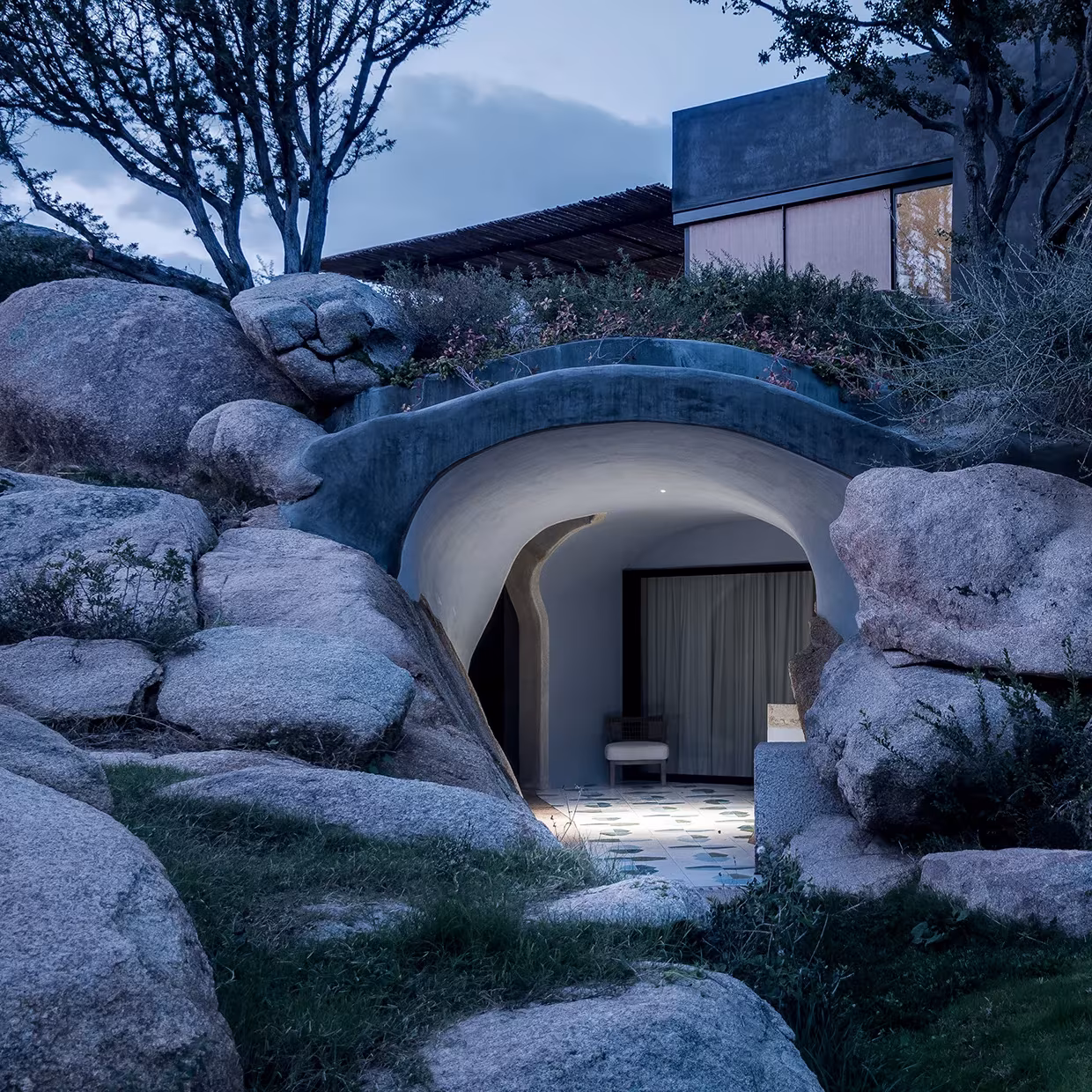 Refugio semienterrado de La Grintosa entre rocas de granito y vegetación, con la casa sobre él al fondo
