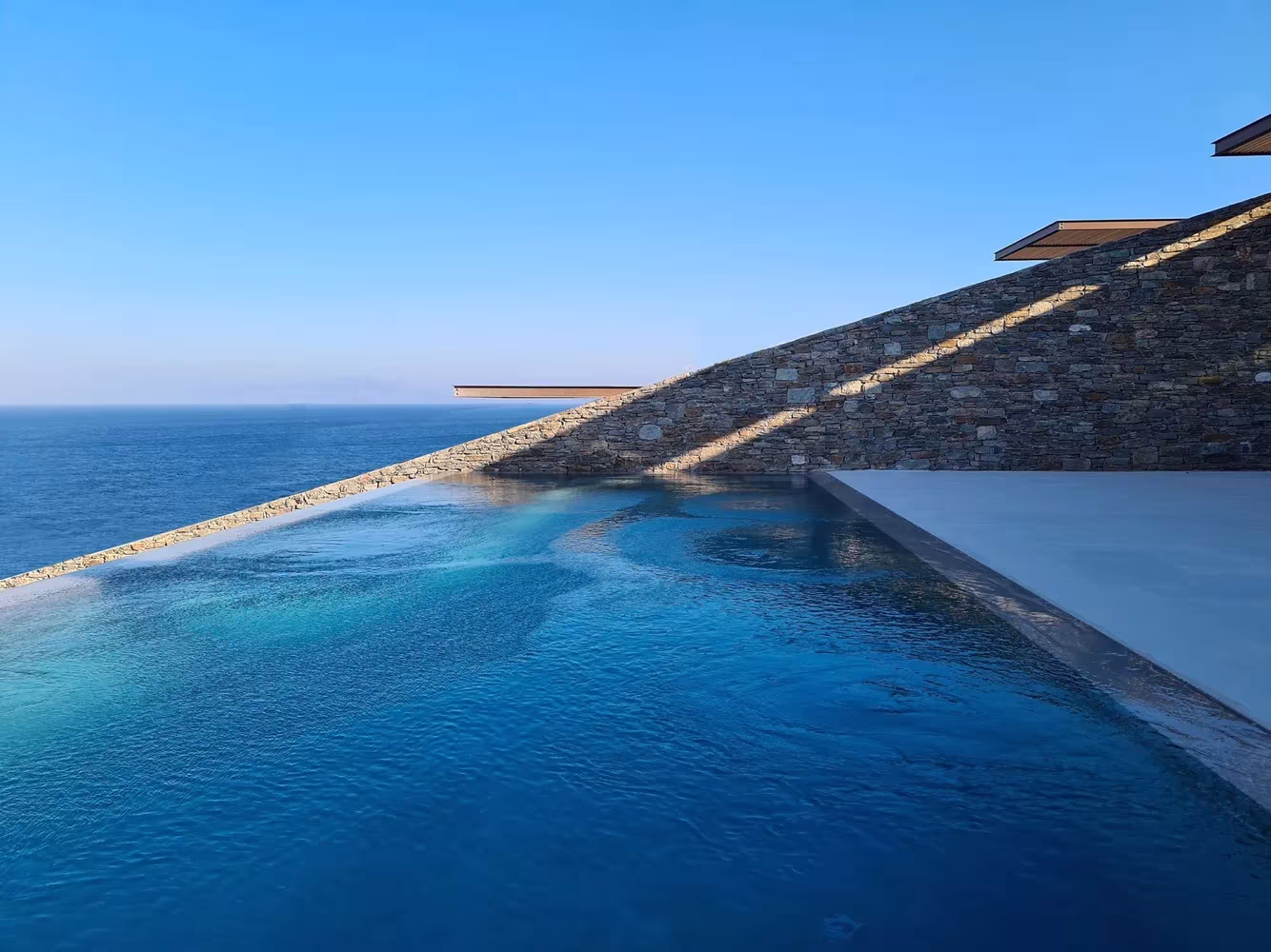 Piscina de borde infinito de la Casa NCaved de Mold Architects sobre el mar Egeo, con muros de piedra en la ladera