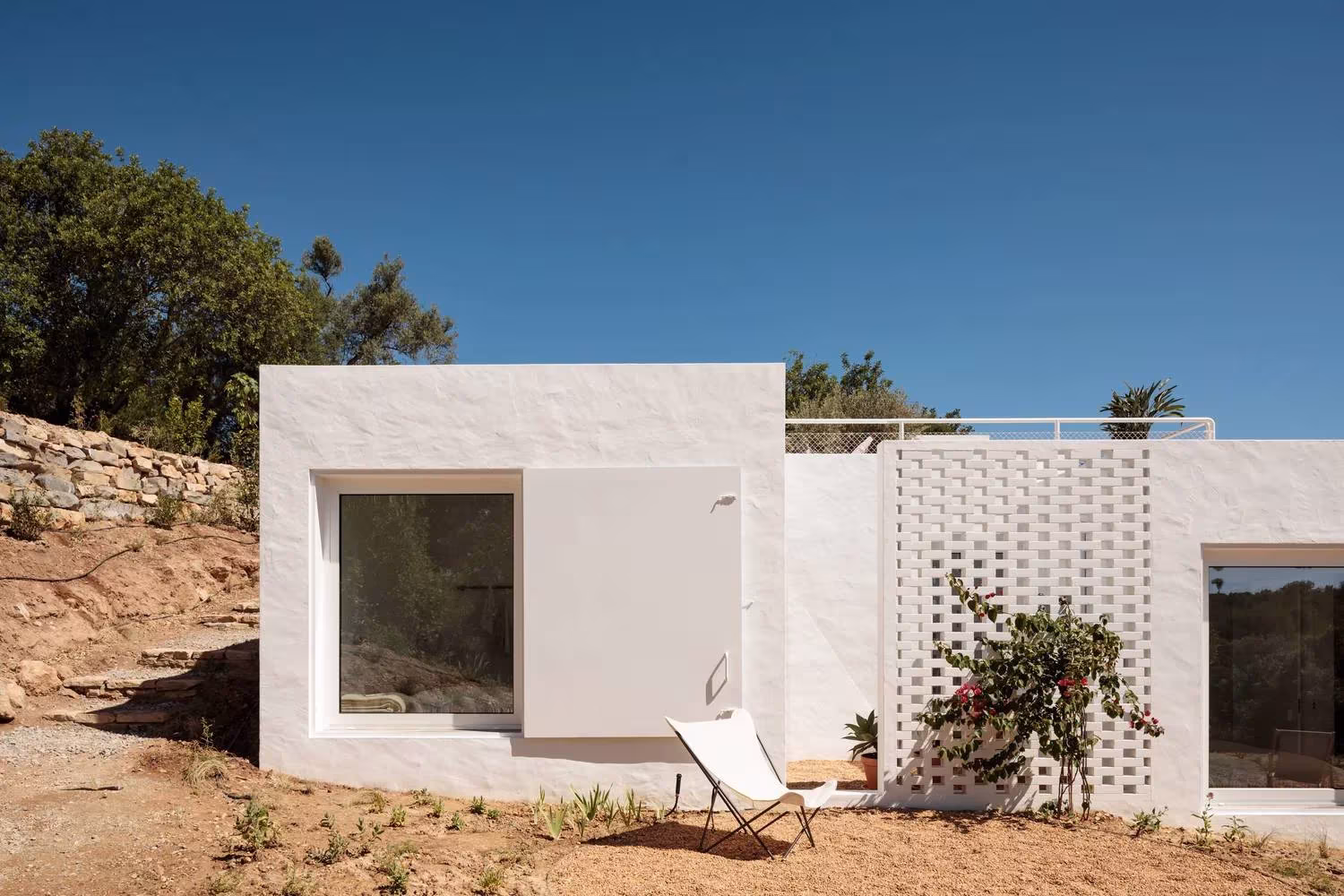 Cuerpo blanco con ventana profunda y celosía cerámica sobre un muro de piedra seca en la Casa UM