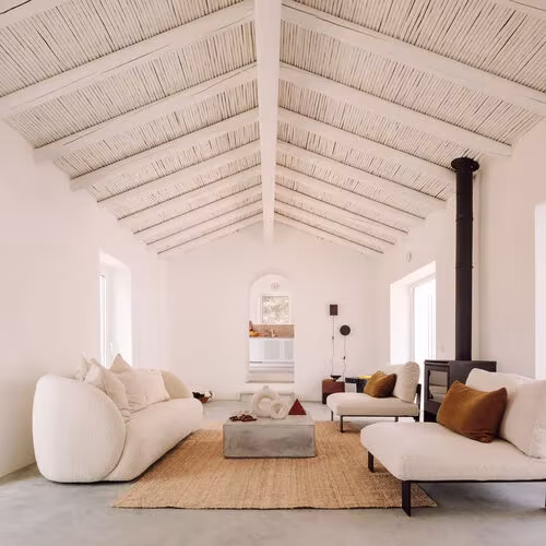 Salón bajo cubierta a dos aguas de cañizo, con sofás de bouclé y estufa negra en la Casa UM