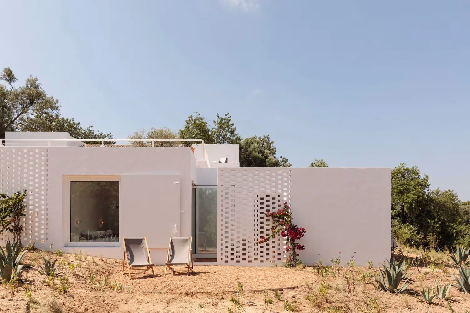 Fachada blanca con celosía de ladrillo hueco y ventana cuadrada entre olivos en la Casa UM