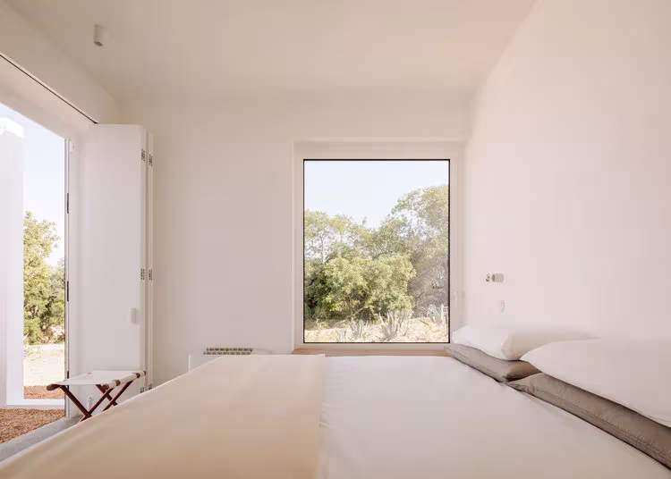Dormitorio blanco con cama baja y ventanas cuadradas abiertas al olivar en la Casa UM